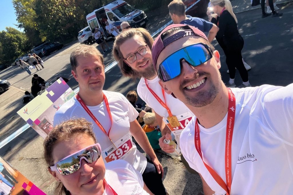 Selfie czworga biegaczy w białych koszulkach Ledwo biegło, z medalami Biegu Medyka na czerwonych smyczach, w słońcu po biegu.